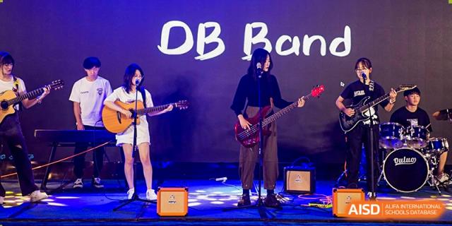 A student band from Milton International School Qingdao performing live on stage with guitars and a bass in front of a dark backdrop with "DB Band" written on it.