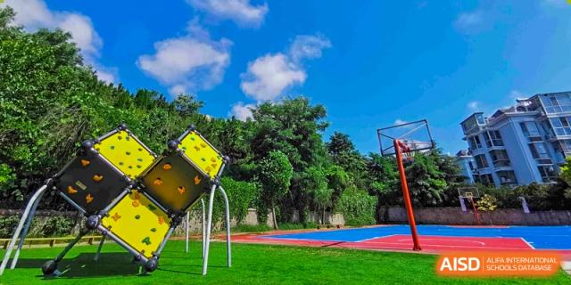 An outdoor playground area at Milton International School Qingdao featuring a colorful sports court and a unique yellow play structure, surrounded by green trees under a blue sky.