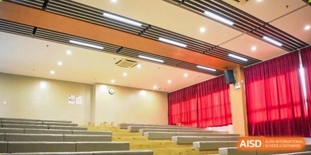 An interior view of a spacious lecture hall or large classroom at Milton International School Qingdao with rows of wooden desks and a stage area with red curtains.