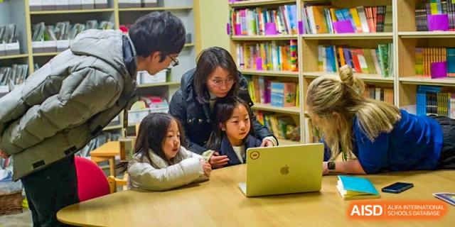 An adult mentor or teacher helping Nanjing International School students with schoolwork on a laptop in a library setting.