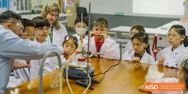 Young Nanjing International School students, wearing lab coats and safety goggles, watch a science demonstration in a classroom.
