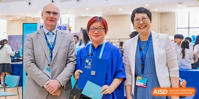 A Nord Anglia Chinese International School Shanghai teacher or staff member smiling and holding a blue folder while interacting with students or colleagues.