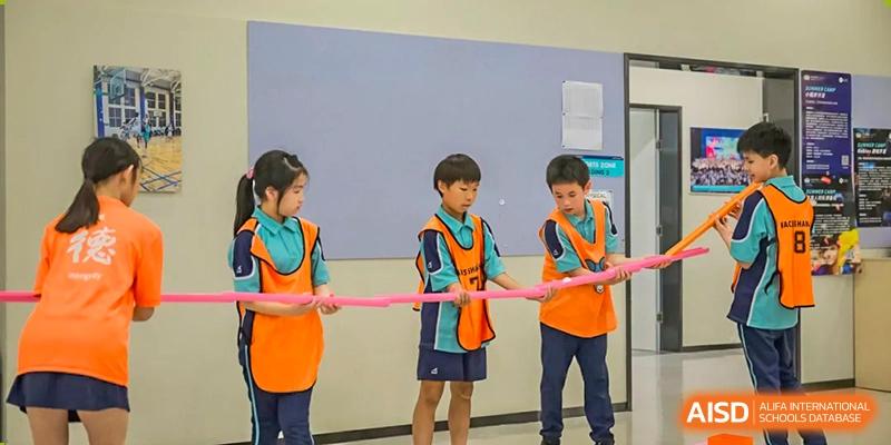 Nord Anglia Chinese International School Shanghai students wearing orange and blue vests holding a pink ribbon for a race or activity indoors.