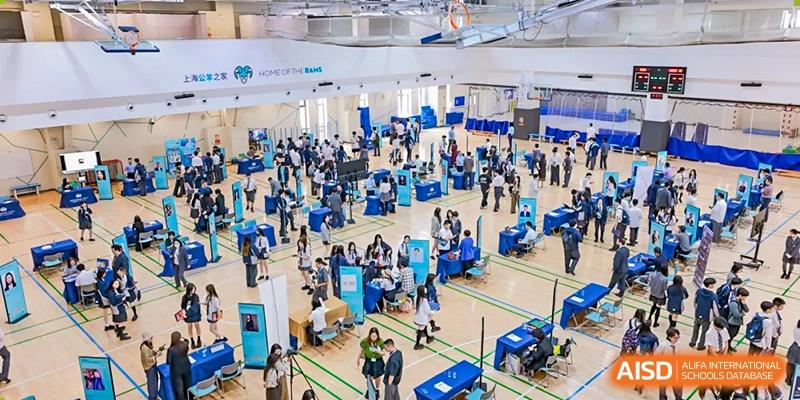 A busy indoor fair or event taking place in a large gymnasium at Nord Anglia Chinese International School Shanghai, with many people and blue tables.