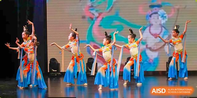 A group of Nord Anglia Chinese International School Shanghai students performing a graceful dance in bright blue and orange costumes on a stage.