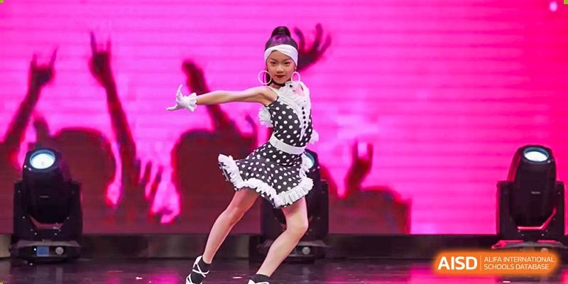 A young Nord Anglia Chinese International School Shanghai student performing a lively dance solo on a pink-lit stage in a black and white polka-dot dress.