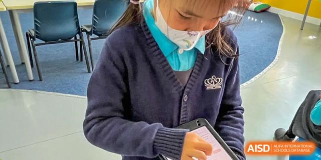 A close-up of a student in a mask and uniform using a tablet or device in a classroom at Nord Anglia International School Shanghai, Pudong.