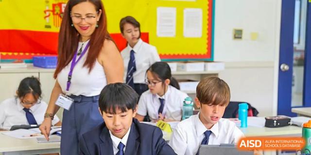 A teacher helping a diverse group of students working on tablets and notebooks in a brightly lit classroom at Nord Anglia International School Shanghai, Pudong.