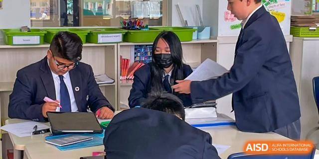 A student in a mask working at a desk while a teacher or classmate stands nearby in a classroom at Nord Anglia International School Shanghai, Pudong.