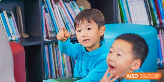 A young boy pointing and smiling while sitting in a classroom or library with bookshelves in the background at Nord Anglia International School Shanghai, Pudong.