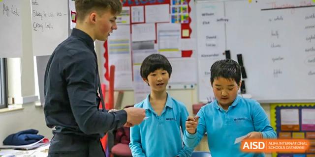 A young male student giving a thumbs up while standing with two other students and a teacher in a classroom at Nord Anglia International School Shanghai, Pudong.