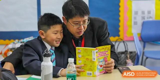 A male teacher in a suit reading a book with two young male students at Nord Anglia International School Shanghai, Pudong.