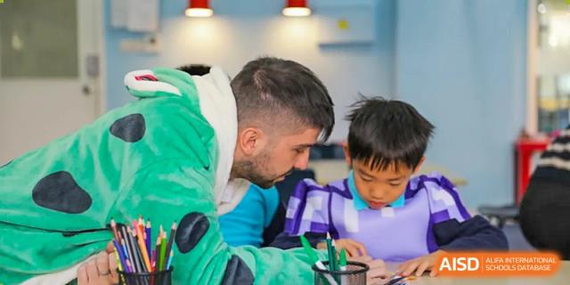A male teacher wearing a costume or playful attire leaning over to assist a young student with a task at Nord Anglia International School Shanghai, Pudong.