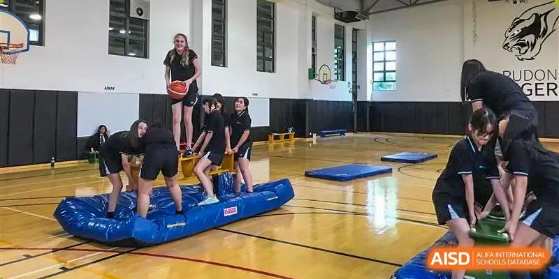 A tall female instructor helping students balance on a blue inflatable obstacle during a gym class at Nord Anglia International School Shanghai, Pudong.