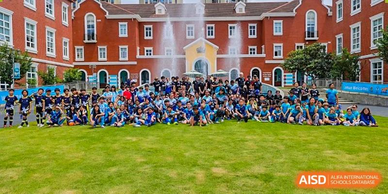A large group of students sitting on the grass in front of a red brick school building with water fountains in the background at Nord Anglia International School Shanghai, Pudong.