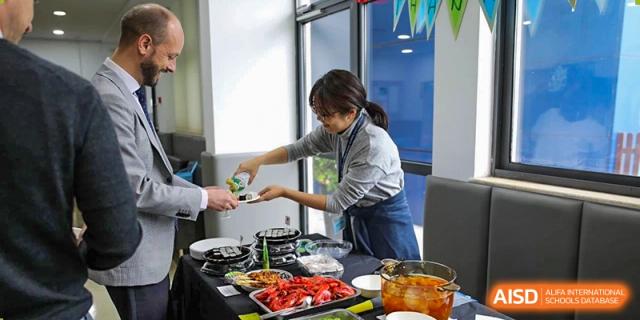 A female cafeteria worker preparing and serving food, including peppers and other cooked items, in the dining hall at Nord Anglia International School Shanghai, Pudong.