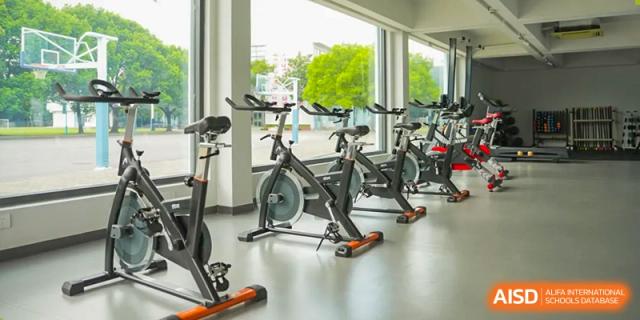 An indoor fitness studio with a row of stationary spin bicycles facing large windows overlooking the outdoor sports courts at Nord Anglia International School Shanghai, Pudong.