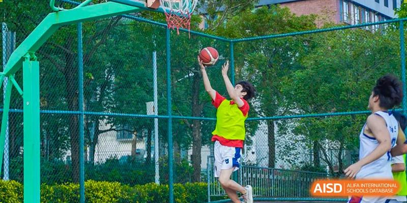 An RDF International School student in a bright vest performs a jump shot while playing basketball outdoors.