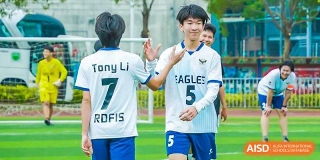Two RDF International School student soccer players, one wearing a jersey with "Tony Li 7 RDFIS" on the back and the other wearing an "EAGLES 5" jersey, high-fiving after a match.