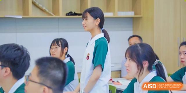 A female RGS Guildford Nanjing student in a white and green polo shirt stands and smiles in a classroom.