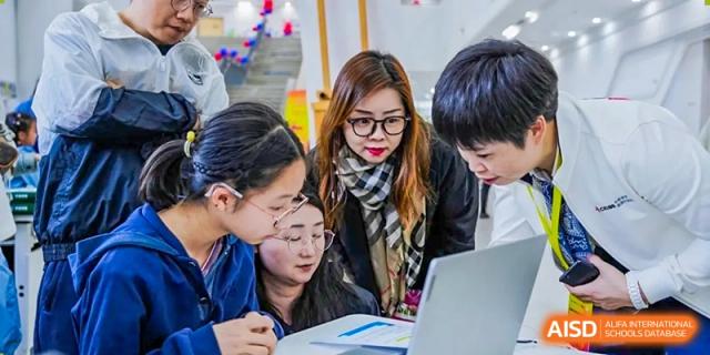 An RGS Guildford Nanjing teacher wearing glasses and a scarf assists a group of students using a laptop.