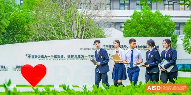 Students in school uniform standing on the campus of RGS Guildford Nanjing, with a large wall displaying text behind them.