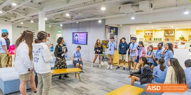Older students stand and listen to a presentation or lecture in a modern, brightly lit common area at Shanghai Community International School.