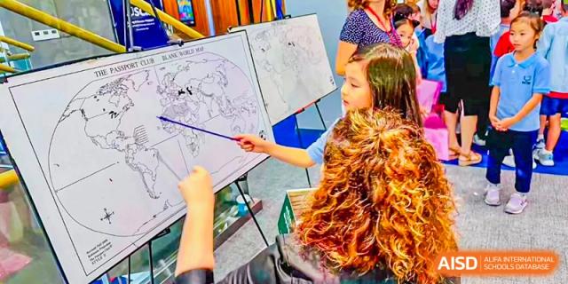 An elementary student points at a world map drawn on an easel, engaging in an interactive lesson with classmates at Shanghai Community International School.
