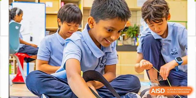 A young male student in a light blue polo shirt smiles while working on a classroom activity with classmates at Shanghai Community International School.