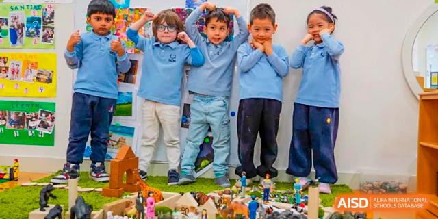 Four young male students in light blue uniforms stand together in front of a small diorama or project display at Shanghai Community International School.