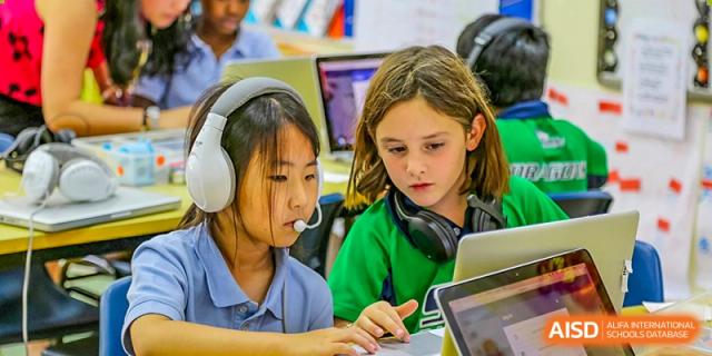 Two female students wearing headsets collaborate on a project using their laptops in a classroom or lab at Shanghai Community International School.