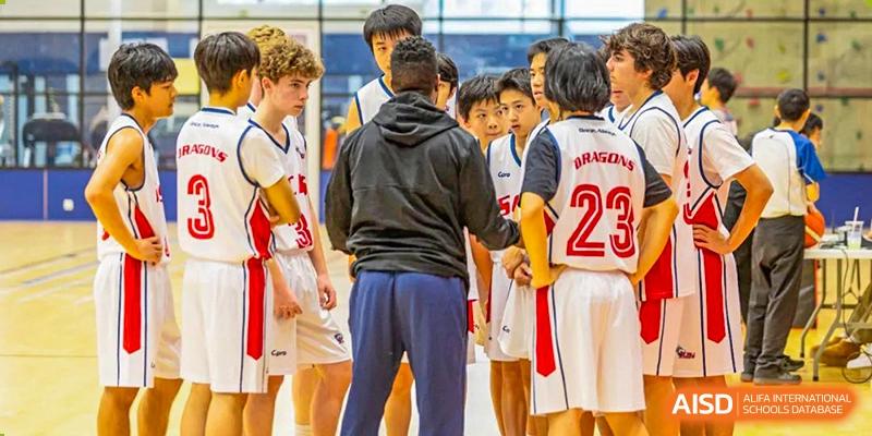 A diverse group of male and female teenage basketball players in red and white uniforms huddle with a coach before a game at Shanghai Community International School.
