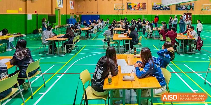 Students are deeply focused and seated at tables taking an exam or participating in a competition in the gymnasium at Shanghai Community International School.