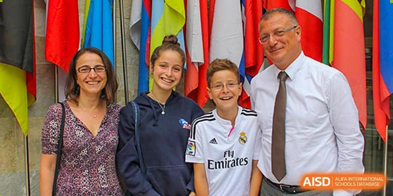 Two smiling students, a girl and a boy wearing a white soccer jersey, pose together in front of a display of international flags at Shanghai Community International School.