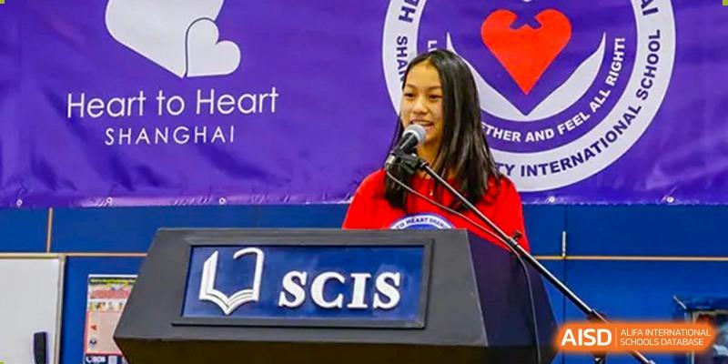A female student in a red shirt gives a speech at a podium with a microphone, with the school logo (SCIS) visible below her, during an event at Shanghai Community International School.