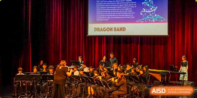 The Dragon Band of Shanghai Community International School performs a piece in a formal auditorium with red curtains.