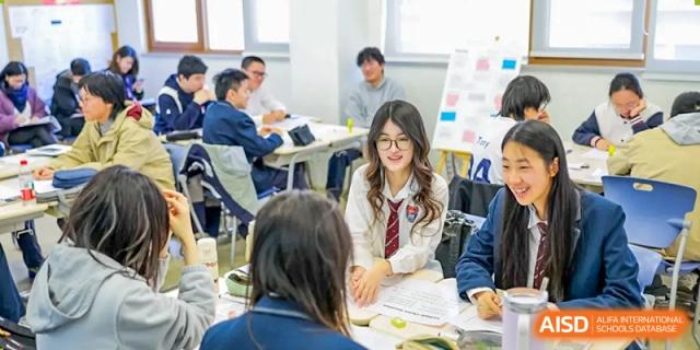 Two female students in uniforms smile at each other while collaborating on a group assignment in a brightly lit classroom at Shanghai DTD Academy.