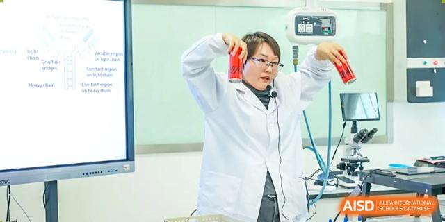 A male student in a lab coat and headset speaks passionately, possibly presenting or teaching, in a classroom at Shanghai DTD Academy.