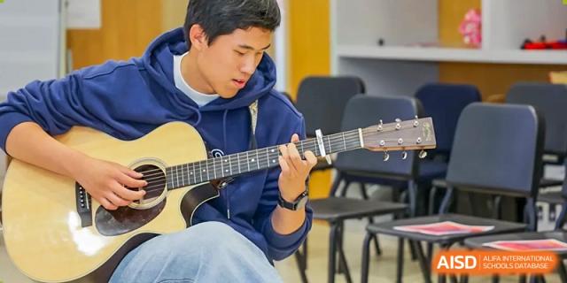 A male student in a blue hoodie practices playing an acoustic guitar in a classroom or rehearsal space at Shanghai DTD Academy.
