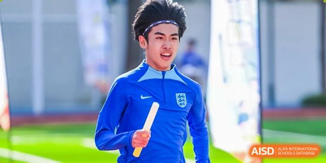 A male student athlete in a blue uniform with an England crest holds a baton, ready to run a relay race for Shanghai DTD Academy.