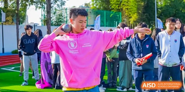 A male student in a bright pink hoodie prepares to throw a shot put or heavy object during a track and field competition at Shanghai DTD Academy.