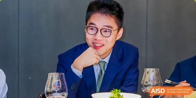 A young man in a suit and tie, smiling and resting his chin on his hand, is seated at a formal event at Shanghai DTD Academy.