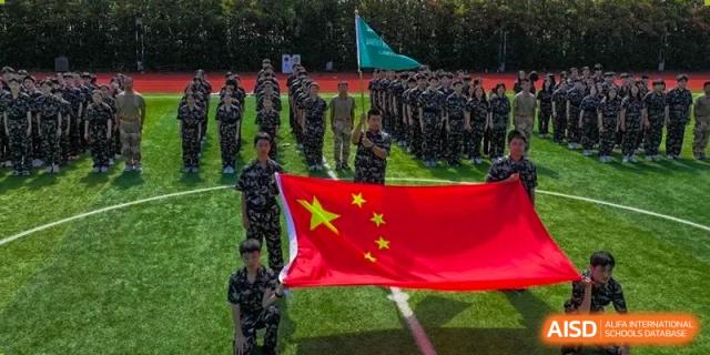 Students or military personnel from Shanghai New Epoch Bilingual School standing in formation on a field during a ceremony, with a large Chinese flag being held in the foreground.