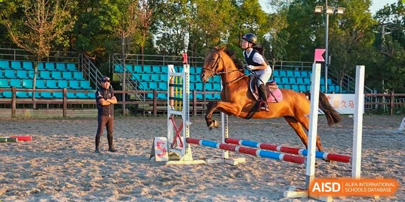 A student from Shanghai New Epoch Bilingual School wearing a helmet and riding a horse over a jump in an outdoor equestrian arena.
