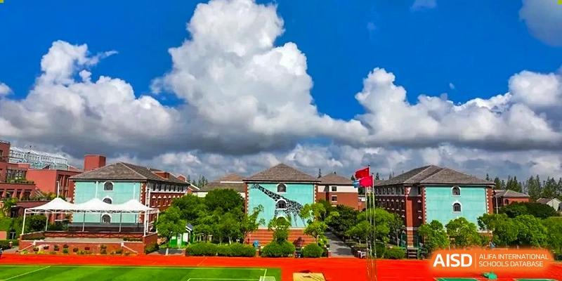An outdoor sports field and school building at Shanghai New Epoch Bilingual School on a sunny day with white clouds, showing the red track and green turf.
