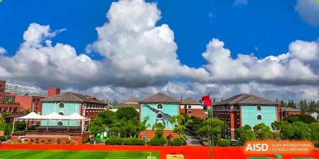 An outdoor sports field and school building at Shanghai New Epoch Bilingual School on a sunny day with white clouds, showing the red track and green turf.