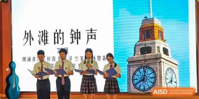 Students in uniform giving a presentation or speech on a stage with a screen displaying Chinese text, at Shanghai United International School, Wanyuan Campus.