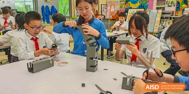 A female student in a blue top carefully working on a hands-on construction or engineering project at her desk at Shanghai United International School, Wanyuan Campus.