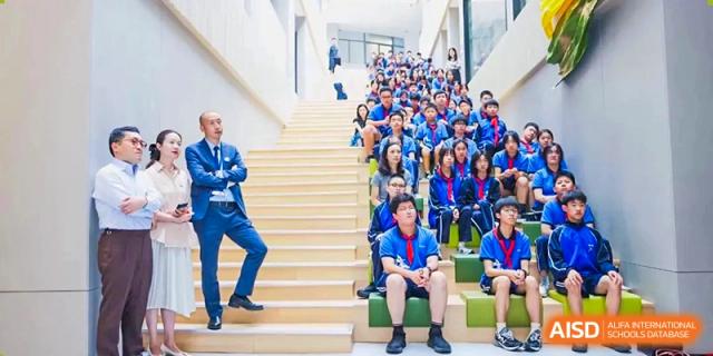 A large group of students sitting on a wide indoor staircase, posing for a group photo or waiting for an event to start, at Shanghai United International School, Wanyuan Campus.