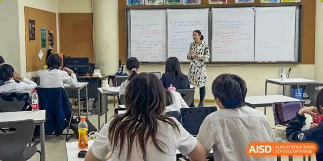 A teacher in a spotted dress giving a lesson to students in a classroom with a whiteboard covered in writing at Shanghai United International School, Wanyuan Campus.